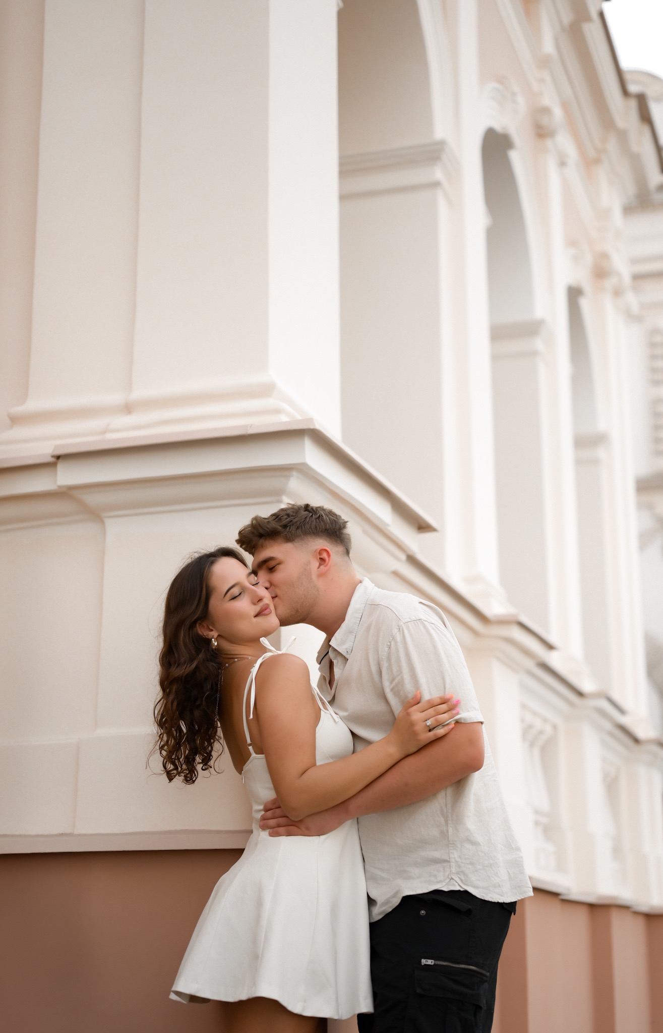 Zakochana para przytulona na tle klasycznej architektury z łukami. Mężczyzna całuje kobietę w policzek. Ona w białej sukience, on w jasnej koszuli.