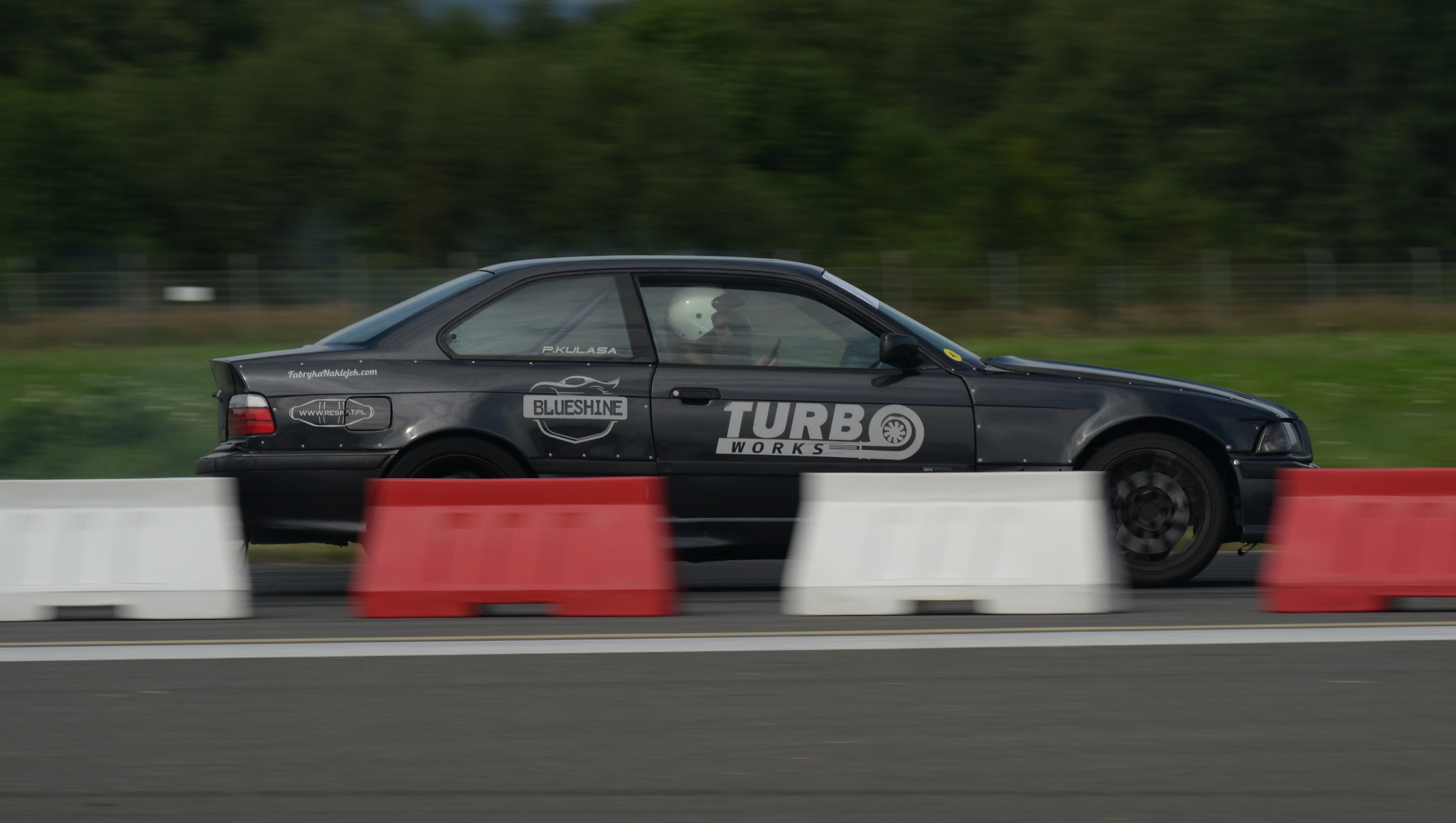 Czarny samochód sportowy BMW E36 z naklejkami sponsorskimi, mknący po torze wyścigowym obok czerwono-białych barier. Kierowca w kasku widoczny wewnątrz.