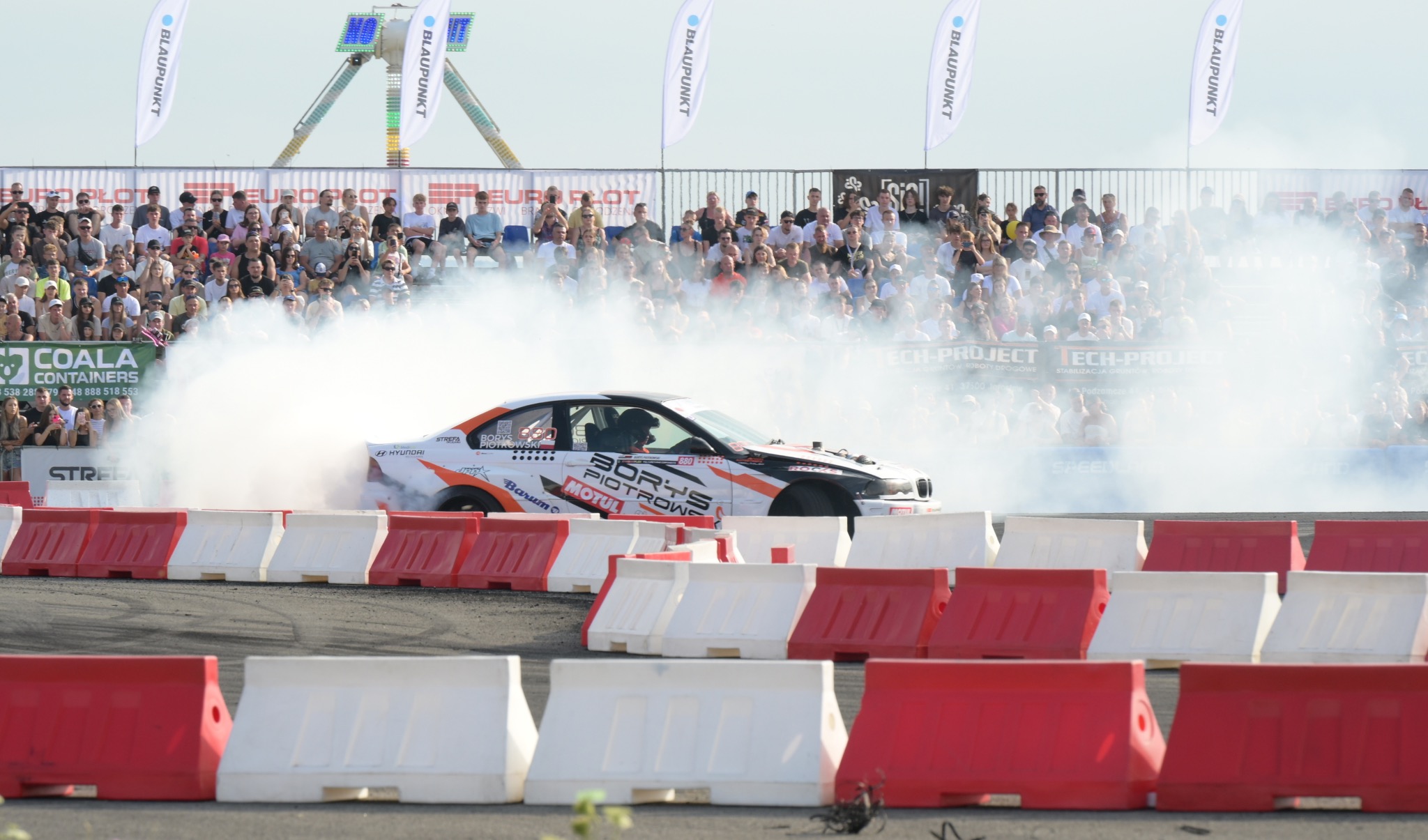 Driftingowy samochód w akcji, otoczony dymem na tle trybun pełnych widzów i banerów reklamowych. Dynamiczne ujęcie z bliska, z widocznymi detalami bolidu.