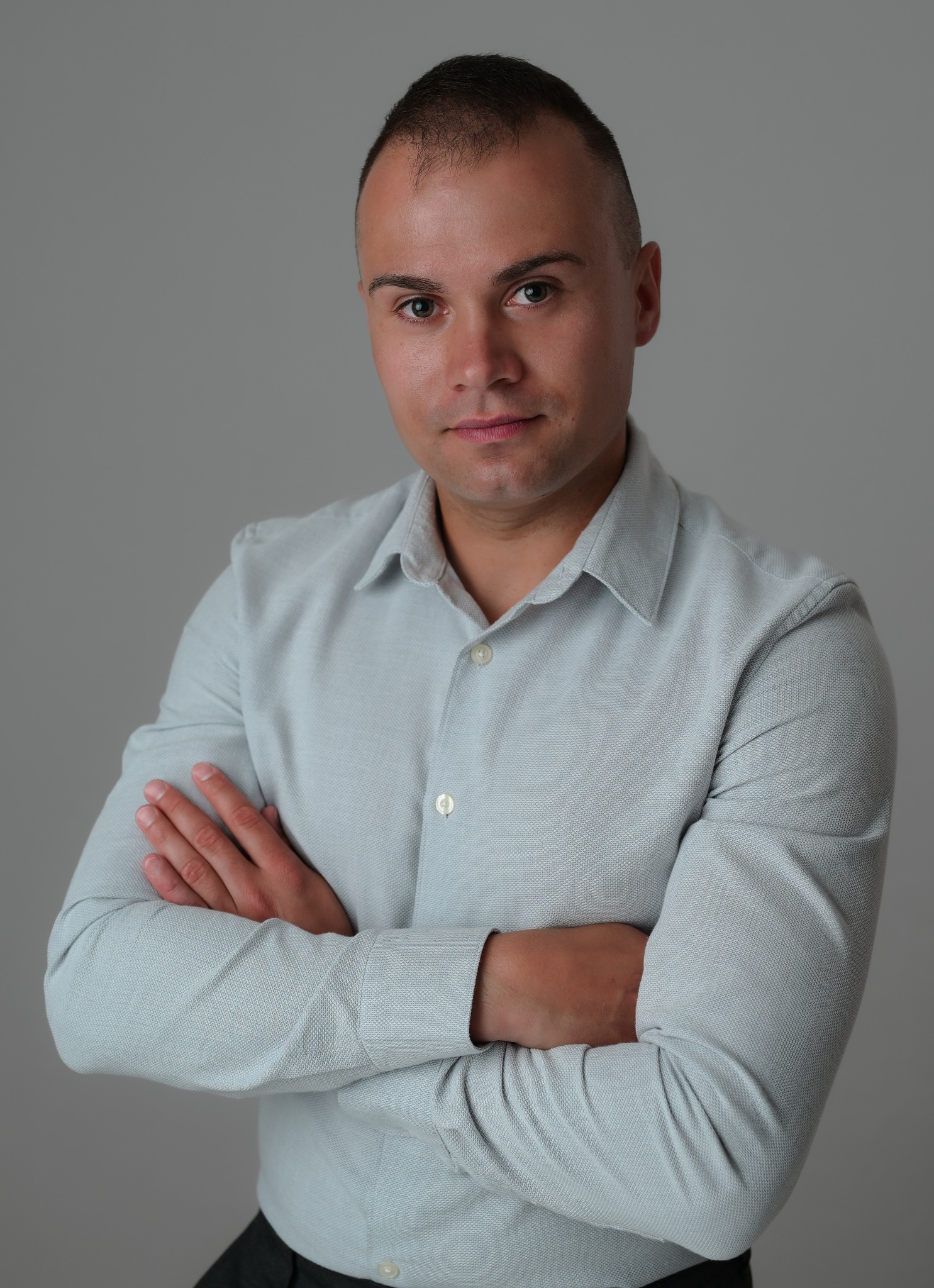 Portret mężczyzny w błękitnej koszuli z założonymi rękoma na szarym tle. Profesjonalny wygląd, ostre światło, biznesowy charakter.
