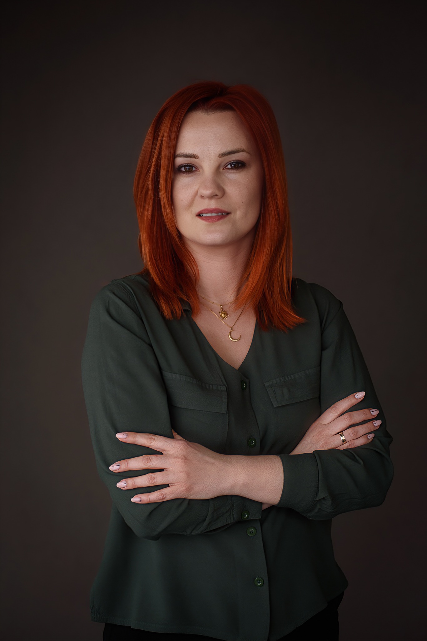 Portret kobiety z rudymi włosami w zielonej koszuli, skrzyżowane ręce, złoty naszyjnik, na ciemnym tle. Uśmiech, profesjonalny wygląd, subtelny makijaż.