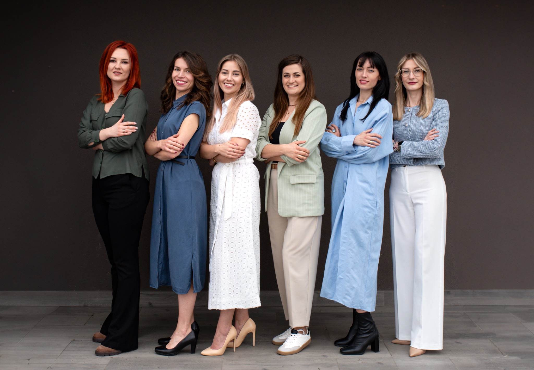 Portret grupowy sześciu uśmiechniętych kobiet w różnorodnych stylizacjach biznesowych na tle szarej ściany. Złożone ręce, pewny wyraz twarzy, naturalne światło.