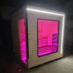 LIFT PACK SPÓŁKA CYWILNA WOJCIECH WADOWSKI, ADRIAN CHMIELARCZYK - Drewniana sauna fińska z przeszklonymi drzwiami i oknem, podświetlona różowym światłem LED, z oświetleniem zewnętrznym. Widok z boku, w ciemnym otoczeniu.