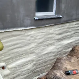 Świeżo nałożona, jasna pianka izolacyjna na fundamencie budynku z dwoma oknami, żółty wąż do aplikacji pianki widoczny po lewej stronie.