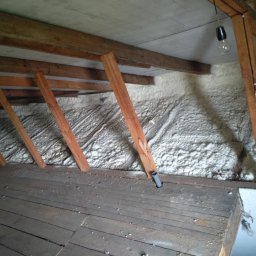 ADRA Izolacje Sp. Z O.O. - Poddasze z drewnianą konstrukcją i ociepleniem pianką PUR na ścianach, widoczna instalacja elektryczna z żarówką. Stare deski podłogowe.