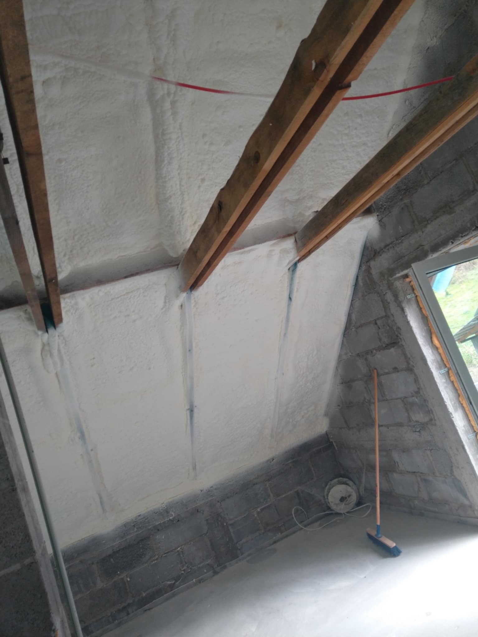 Poddasze po natrysku pianki PUR, widoczne drewniane belki i fragment okna. Ściany z szarych bloczków. Na podłodze szczotka.