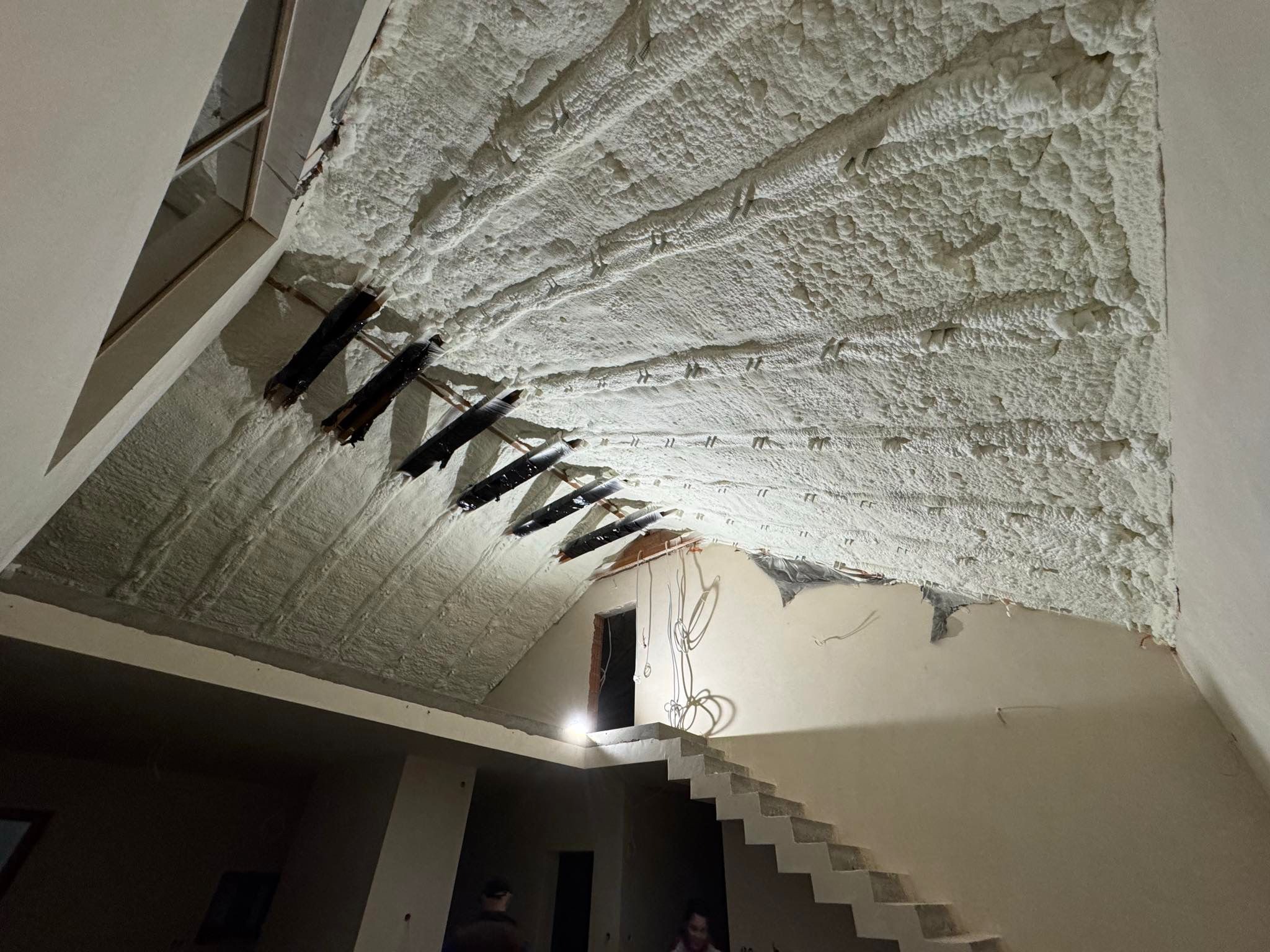 Poddasze po ociepleniu pianką PUR, widoczne drewniane elementy konstrukcyjne. Biała, nierówna powierzchnia izolacji, schody w tle. Detal wykończenia.