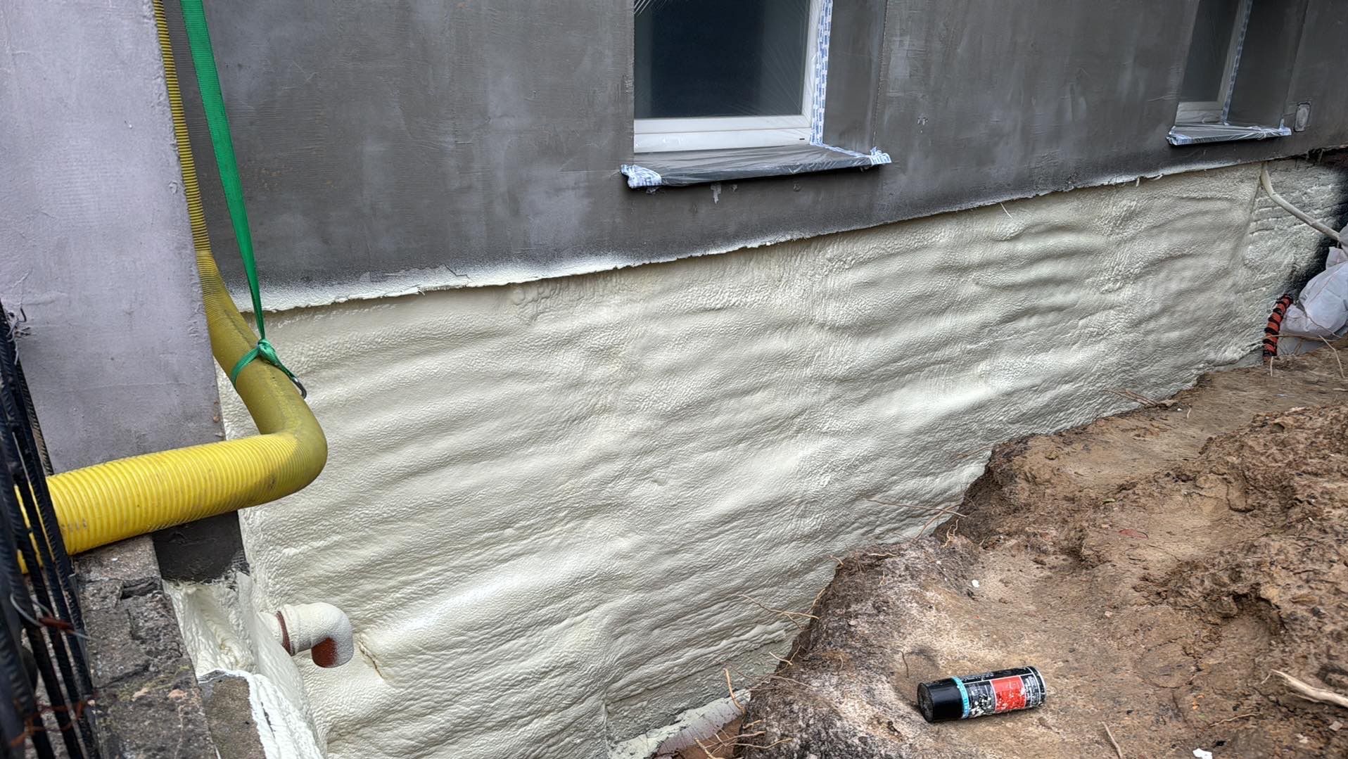 Świeżo nałożona, jasna pianka izolacyjna na fundamencie budynku z dwoma oknami, żółty wąż do aplikacji pianki widoczny po lewej stronie.