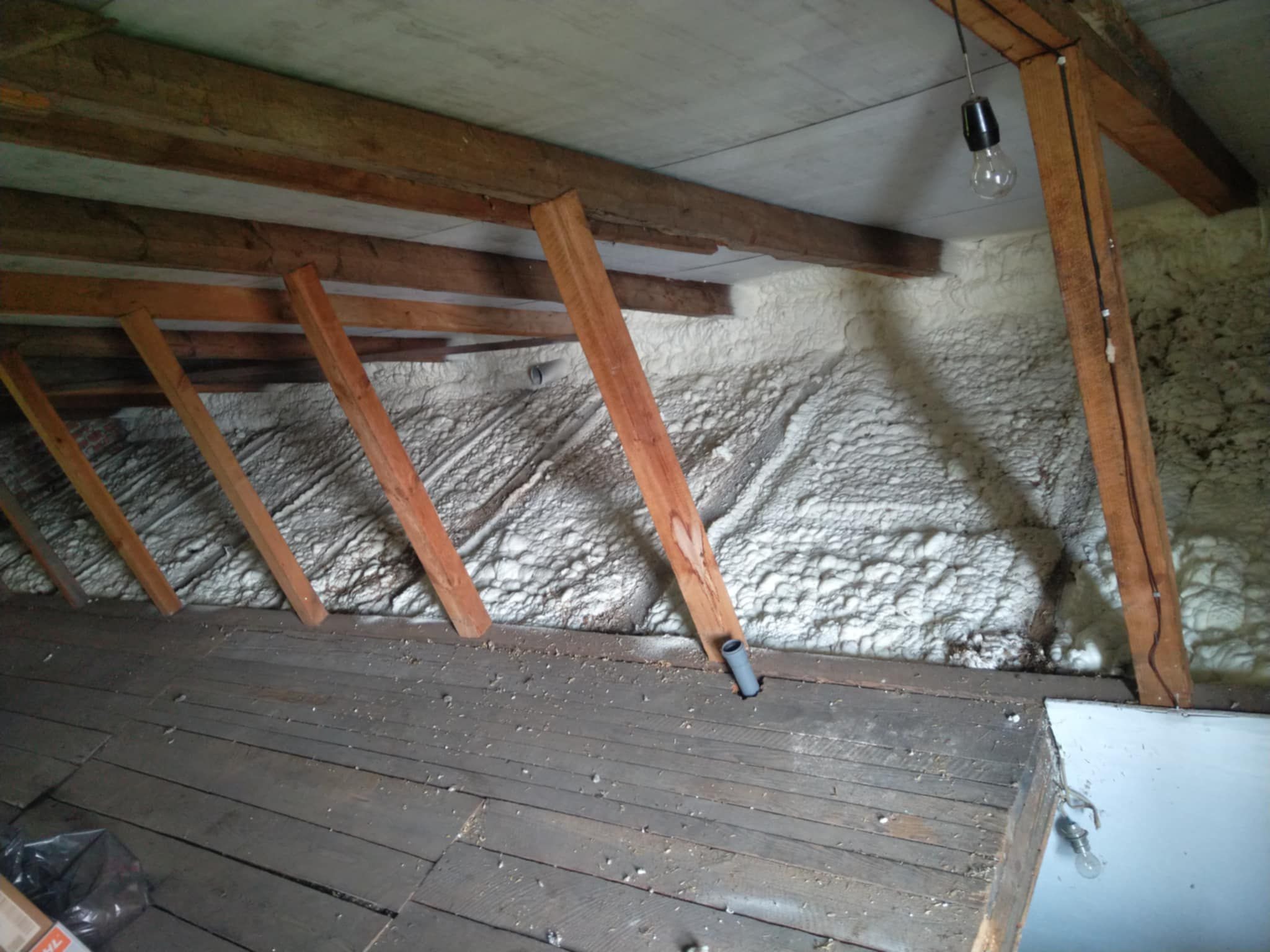 Poddasze z drewnianą konstrukcją i ociepleniem pianką PUR na ścianach, widoczna instalacja elektryczna z żarówką. Stare deski podłogowe.