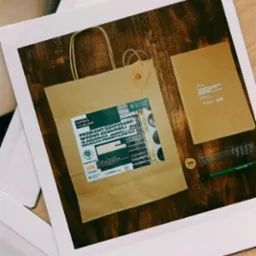 Brązowa torba papierowa z nadrukiem 'Problemy gospodarki wodno-ściekowej' leży na drewnianym stole obok notatnika, prob&oacute;wki i długopisu. Całość sfotografowana w stylu polaroid.