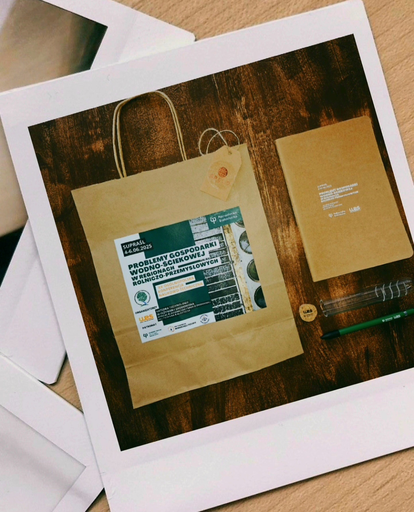 Brązowa torba papierowa z nadrukiem 'Problemy gospodarki wodno-ściekowej' leży na drewnianym stole obok notatnika, probówki i długopisu. Całość sfotografowana w stylu polaroid.