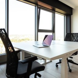 Szopena 35C Sp. z o.o. - Jasne biuro z regulowanym biurkiem, laptopem i ergonomicznymi krzesłami na kółkach, widok na miasto przez duże okna.