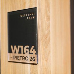 Szopena 35C Sp. z o.o. - Oznakowanie biura: tabliczka z nazwą 'Olszynki Park', numerem W164 i informacją o piętrze 26, zamocowana na drewnianej ścianie.