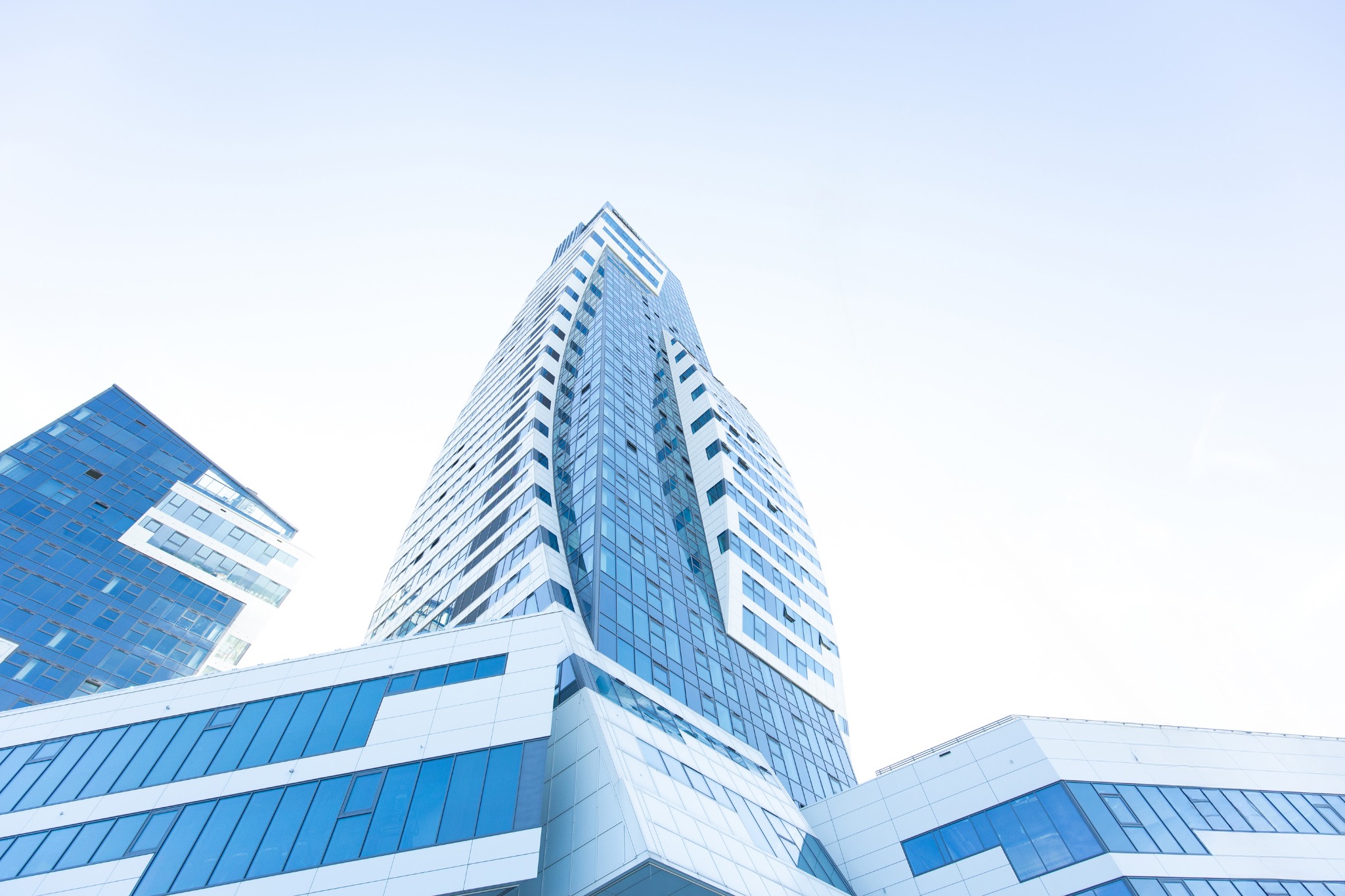 Nowoczesny biurowiec z niebieskimi szklanymi fasadami i białymi elementami architektonicznymi, widziany z dołu pod kątem, na tle jasnego nieba.