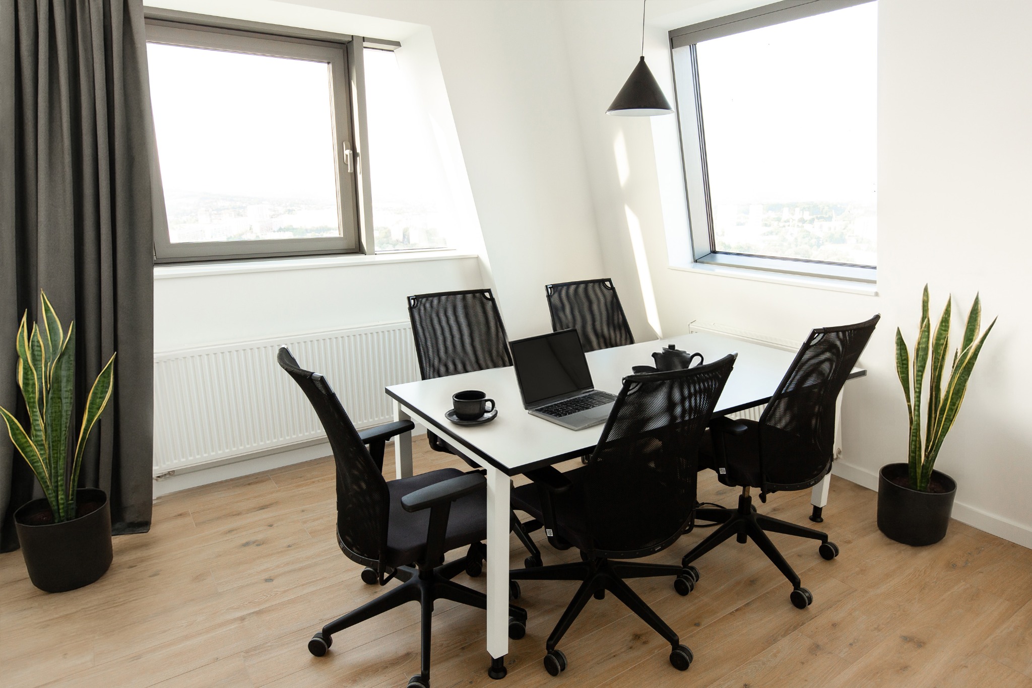 Jasne biuro z oknami, białym stołem, czarnymi krzesłami, laptopem i roślinami doniczkowymi. Minimalistyczny design, drewniana podłoga, nowoczesne oświetlenie.