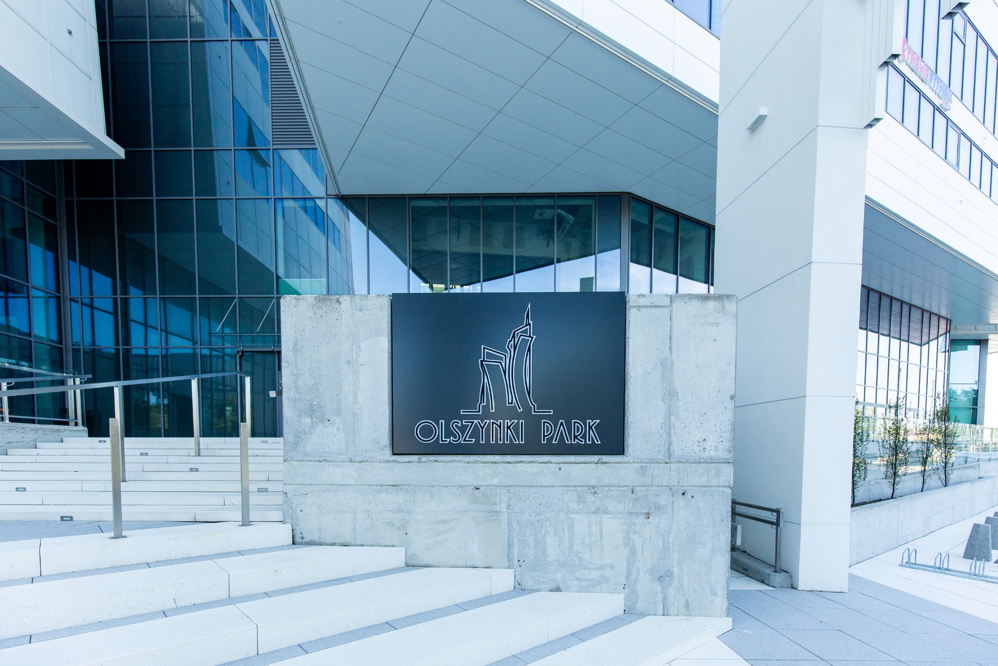Elegancki budynek biurowy Olszynki Park z nowoczesnym logo na betonowym cokole i schodami. Minimalistyczna architektura w jasnej kolorystyce.