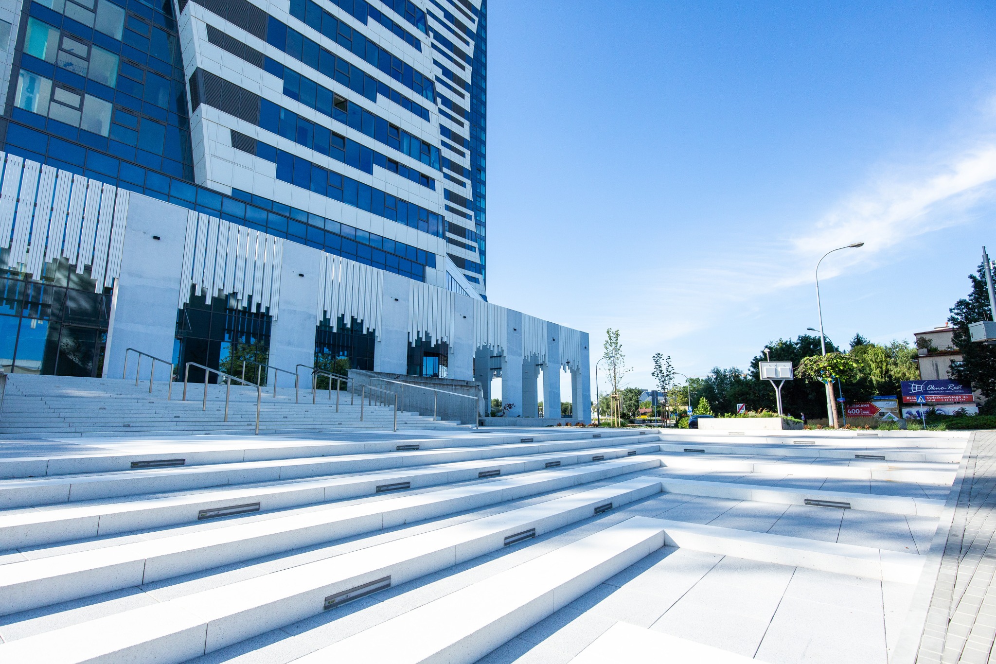 Nowoczesny budynek biurowy z niebieskimi oknami i białymi elementami fasady, widziany z dołu, z szerokimi, białymi schodami prowadzącymi do wejścia.