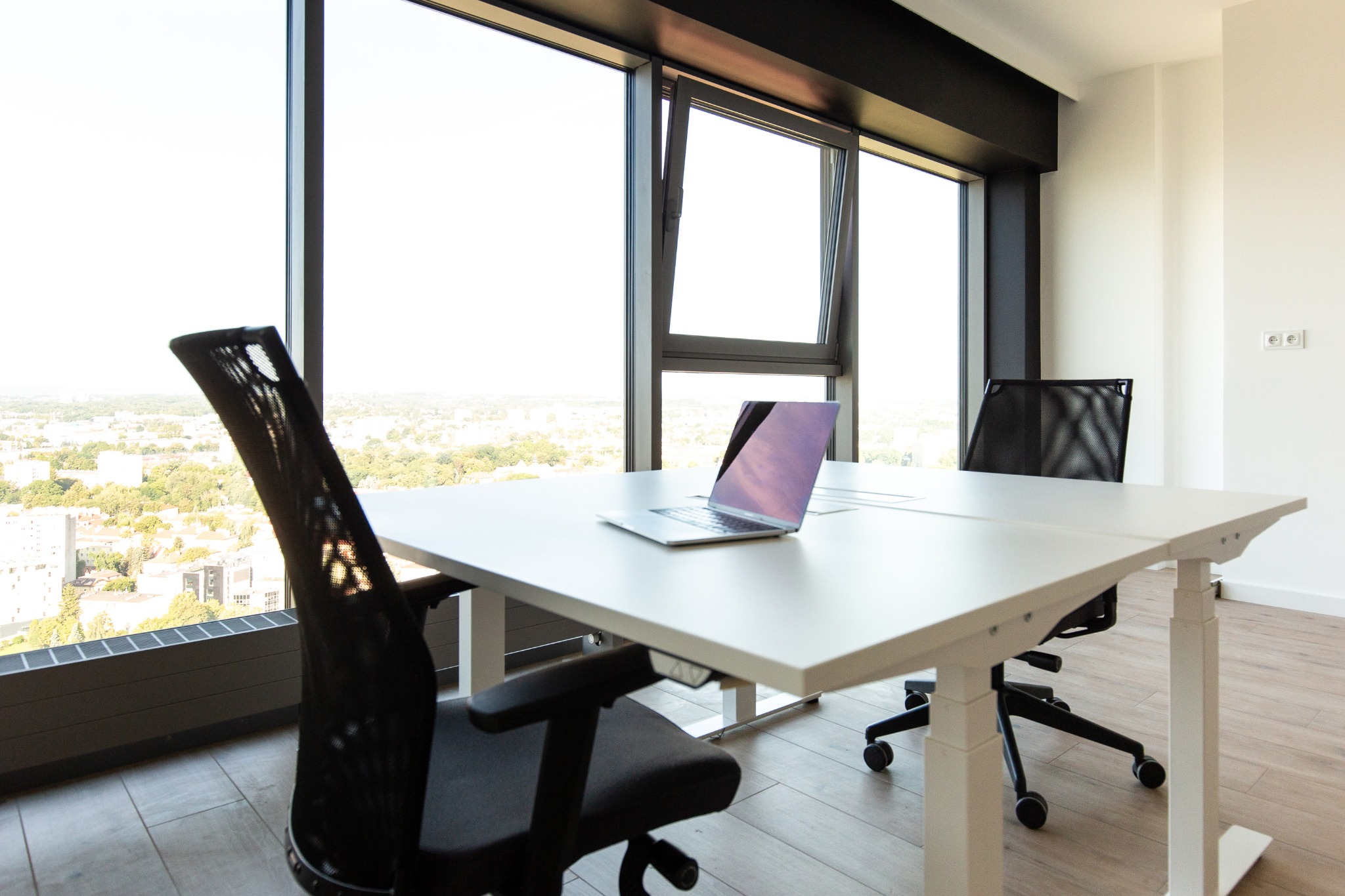 Jasne biuro z regulowanym biurkiem, laptopem i ergonomicznymi krzesłami na kółkach, widok na miasto przez duże okna.