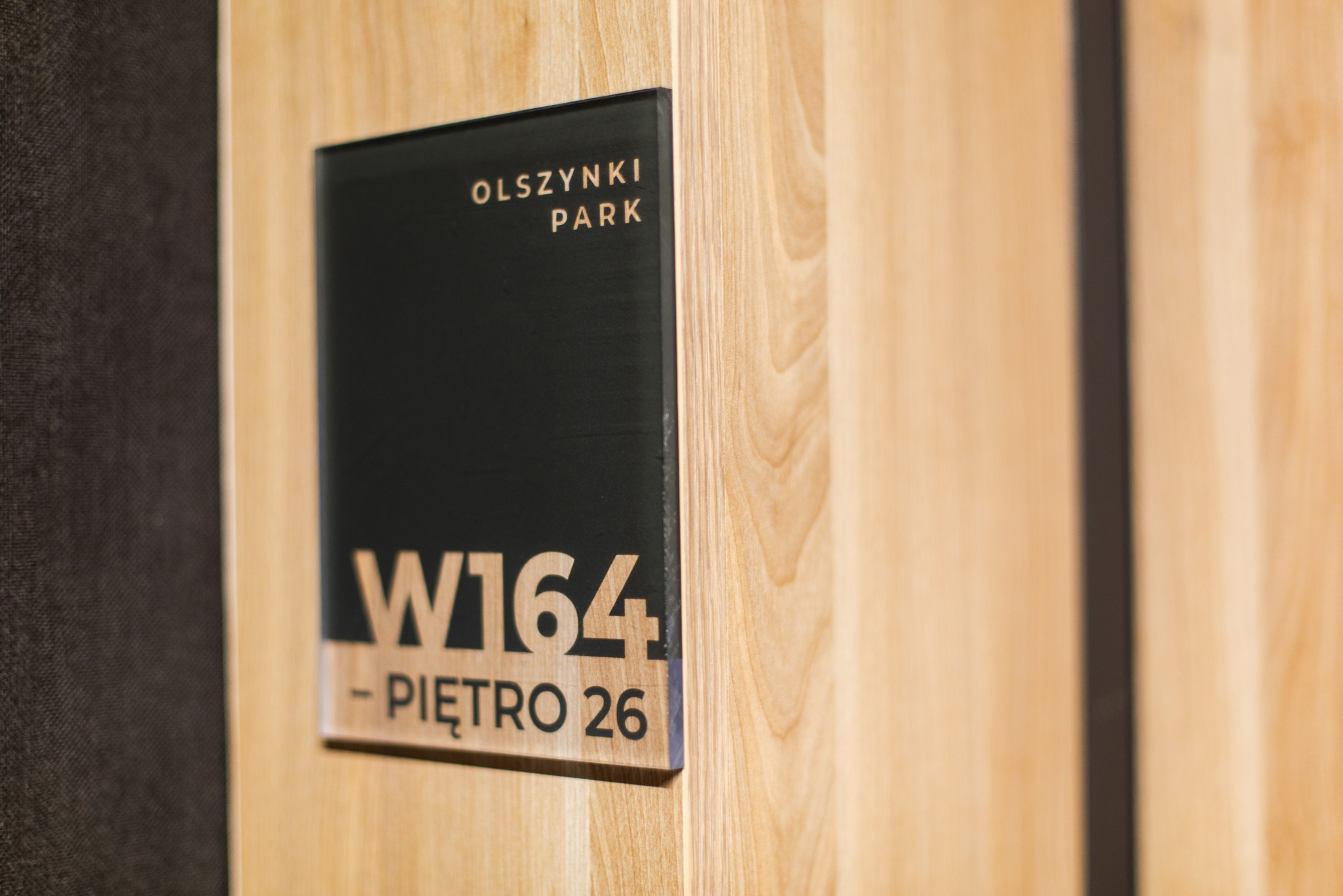Oznakowanie biura: tabliczka z nazwą 'Olszynki Park', numerem W164 i informacją o piętrze 26, zamocowana na drewnianej ścianie.