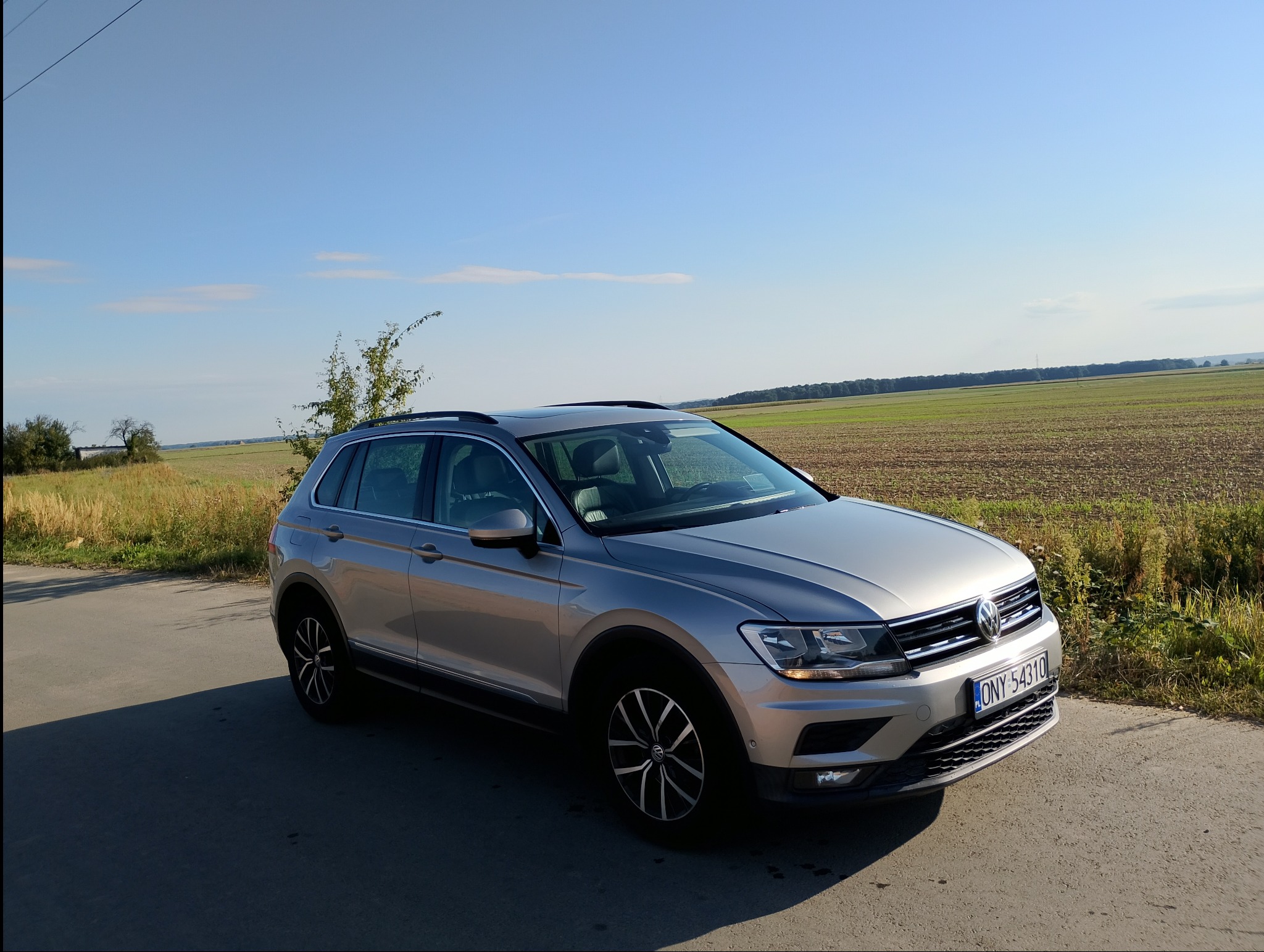Srebrny SUV Volkswagen Tiguan na tle pola i błękitnego nieba. Auto z polską tablicą rejestracyjną, zaparkowane na poboczu drogi.