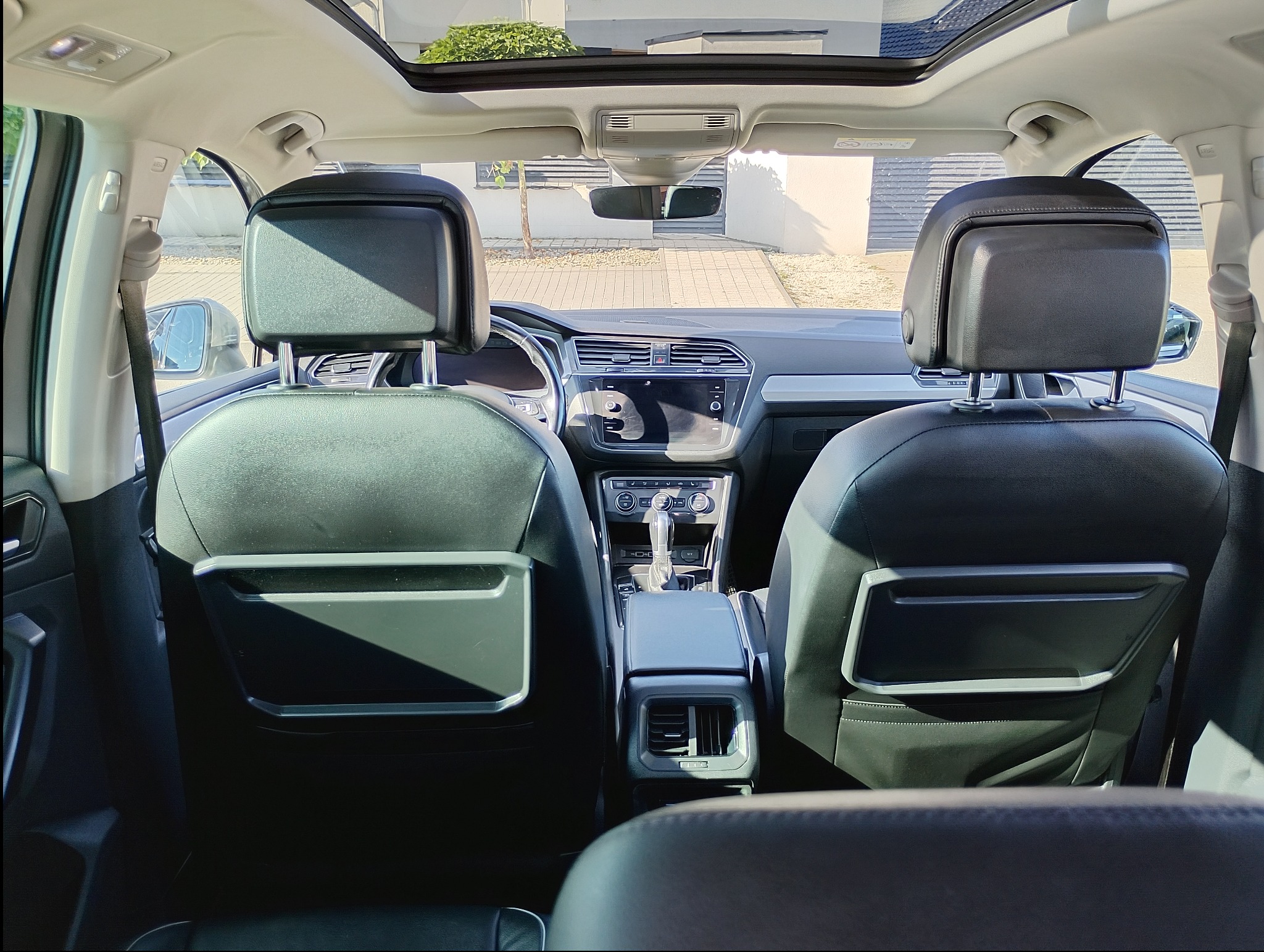 Wnętrze samochodu z czarną skórzaną tapicerką, widok z tylnych siedzeń na deskę rozdzielczą i otwierany szklany dach, jasne otoczenie.