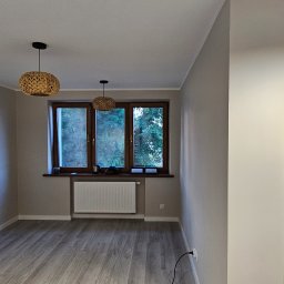 Glądys usługi remontowo budowlane - Jasne wnętrze po remoncie: panele podłogowe, okno z widokiem na zieleń, dwa plecione abażury lamp. Minimalistyczny styl, świeżo odmalowane ściany.