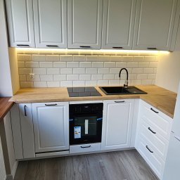 Glądys usługi remontowo budowlane - Biała kuchnia z drewnianym blatem i czarnymi uchwytami, płytki w stylu metro, oświetlenie podszafkowe. Nowoczesny i minimalistyczny design, widok na piekarnik.
