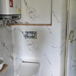 Glądys usługi remontowo budowlane - Biała łazienka z marmurowymi płytkami, widoczny podtynkowy stelaż WC, bojler i kabina prysznicowa z złotymi detalami. Minimalistyczny design i eleganckie wykończenie.