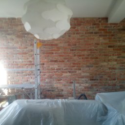 Glądys usługi remontowo budowlane - Murowana ściana z cegły po renowacji, zabezpieczona folią malarską, widoczna drabina i lampa sufitowa. Detal wnętrza po pracach wykończeniowych.