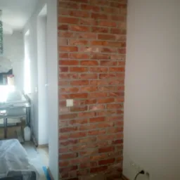Ściana z cegły klinkierowej w jasnym wnętrzu, z widocznym włącznikiem światła i fragmentem umeblowania. Realizacja wykończenia z użyciem cegły dekoracyjnej.