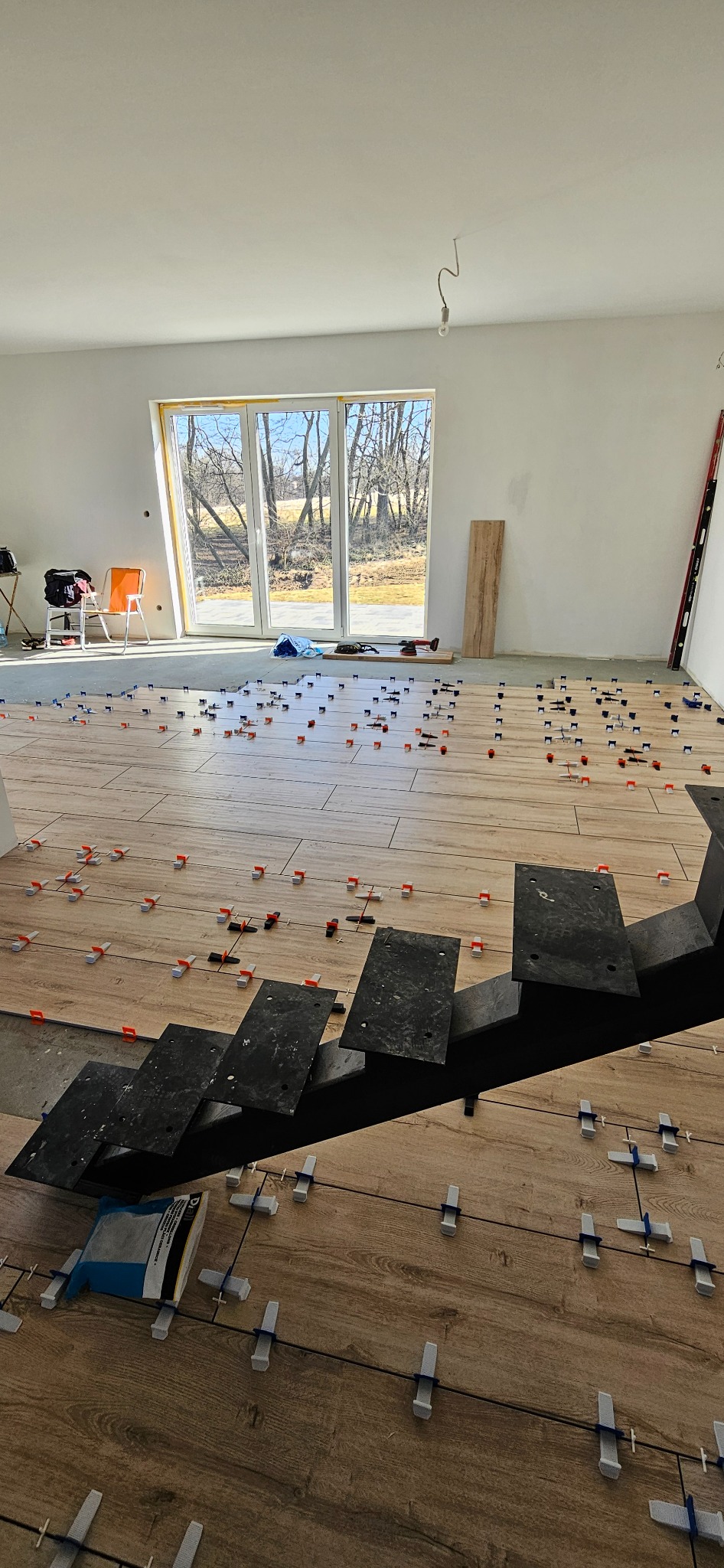 Układanie płytek imitujących drewno w jasnym wnętrzu. Widoczne klipsy i poziomica. Częściowo ułożona podłoga z płytek przy schodach.