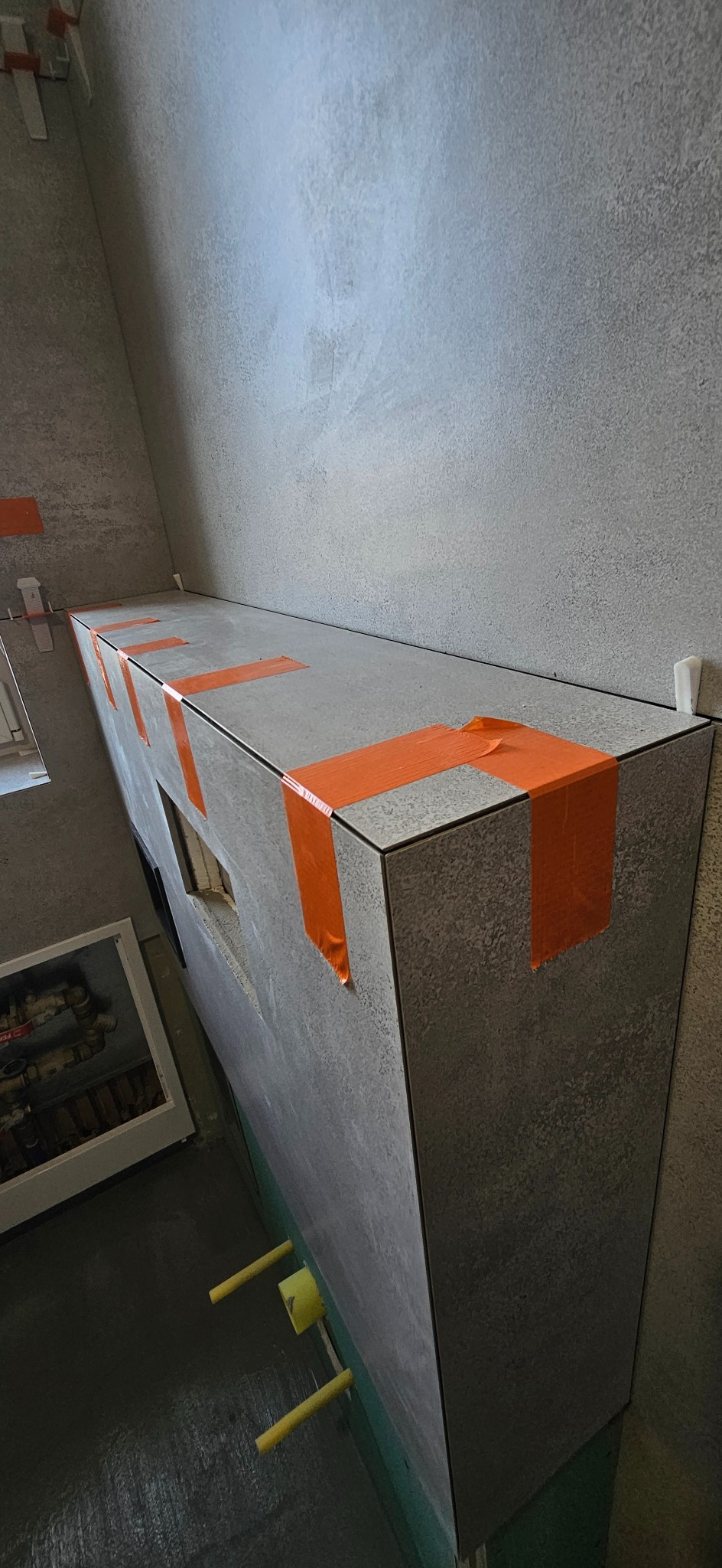 Obudowa z płytek imitujących kamień, zabezpieczona pomarańczową taśmą, w trakcie prac wykończeniowych. Widoczne rury i fragment okna. Szara ściana w tle.