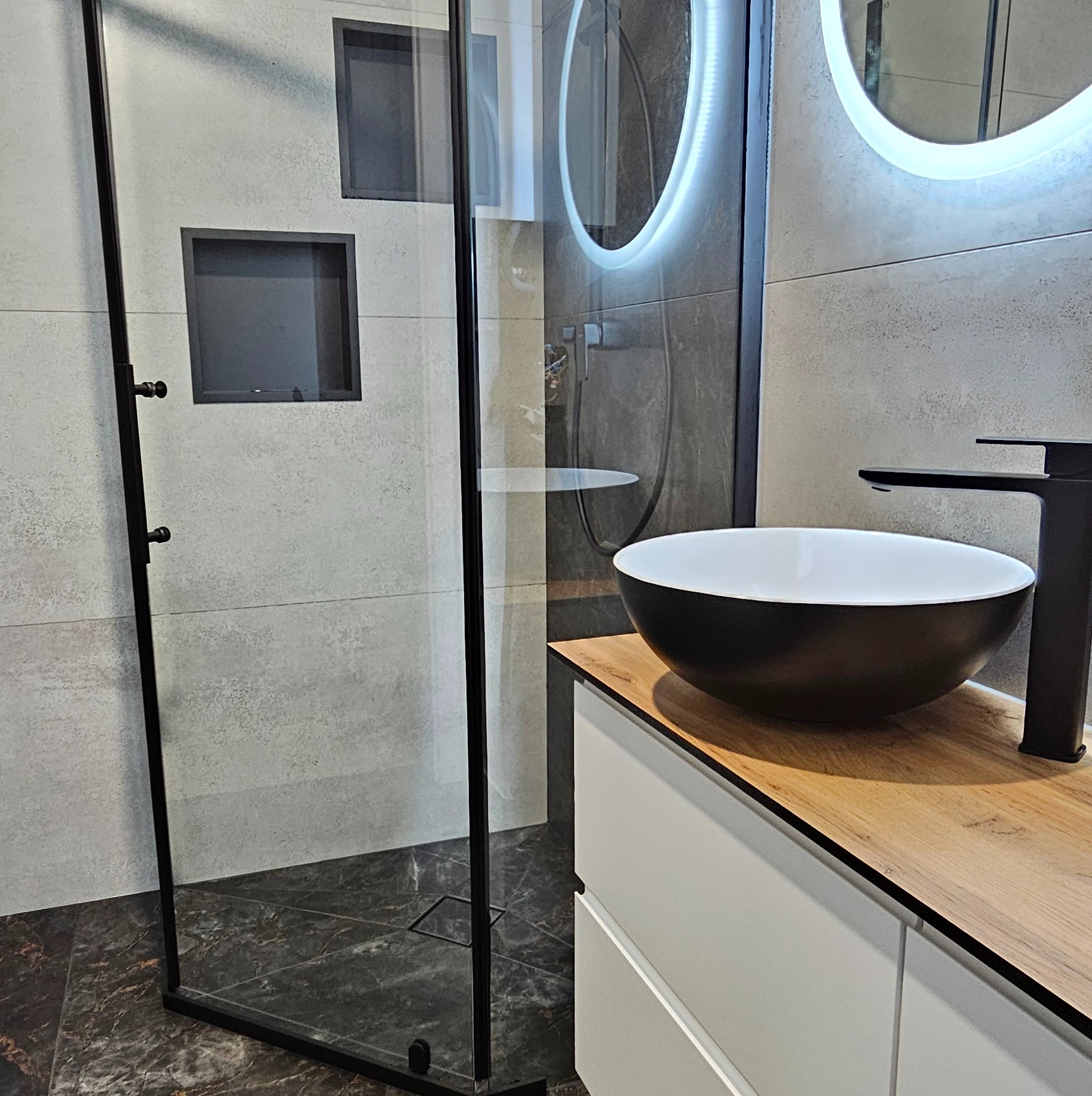 Nowoczesna łazienka: kabina prysznicowa typu walk-in z czarnymi profilami, umywalka nablatowa i podświetlane lustro. Minimalistyczny design, szare płytki i drewniany blat.