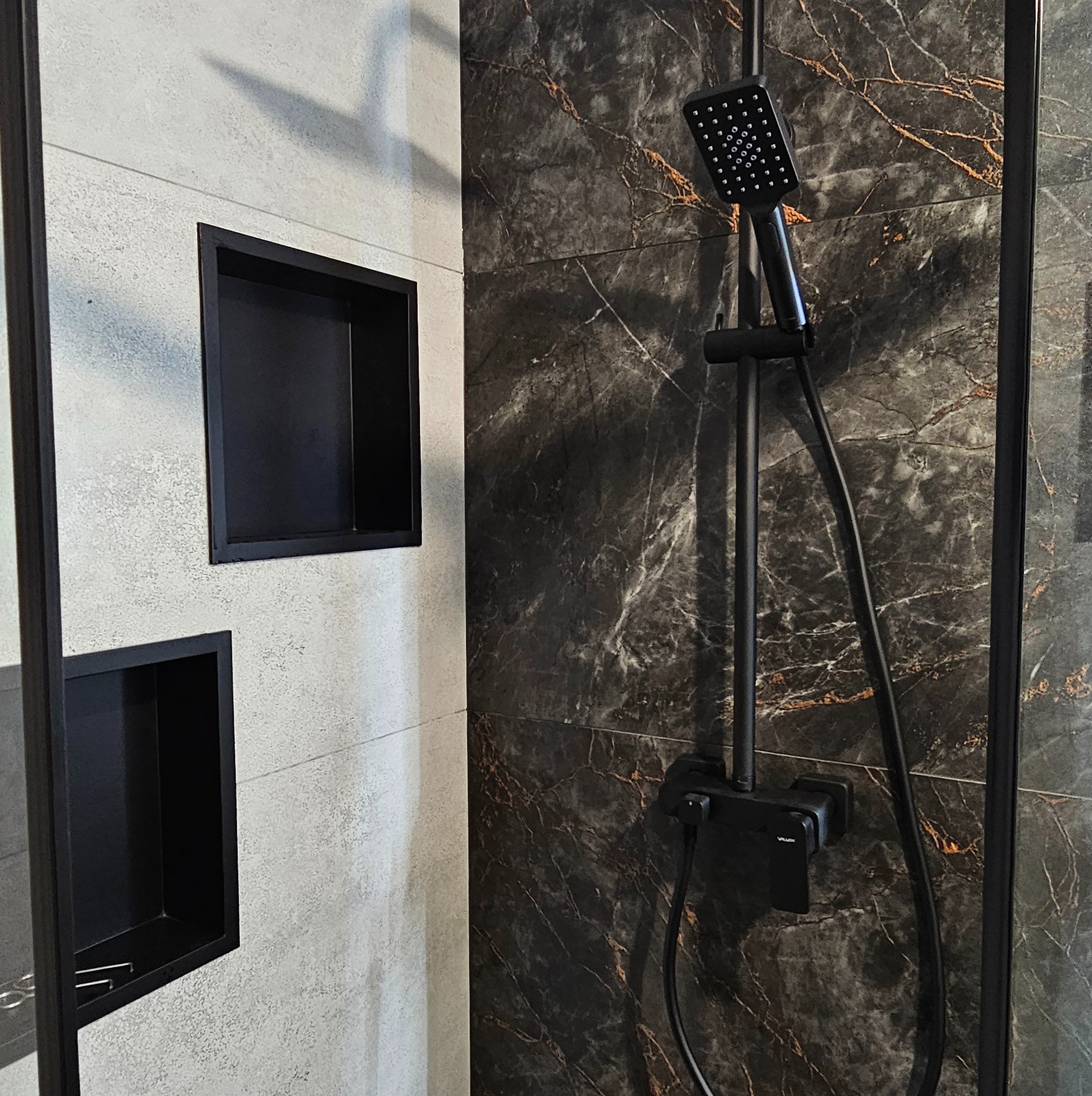 Nowoczesna łazienka z czarną armaturą i wnękami w ścianie wyłożonej szarymi i marmurkowymi płytkami. Minimalistyczny design, eleganckie wykończenie.