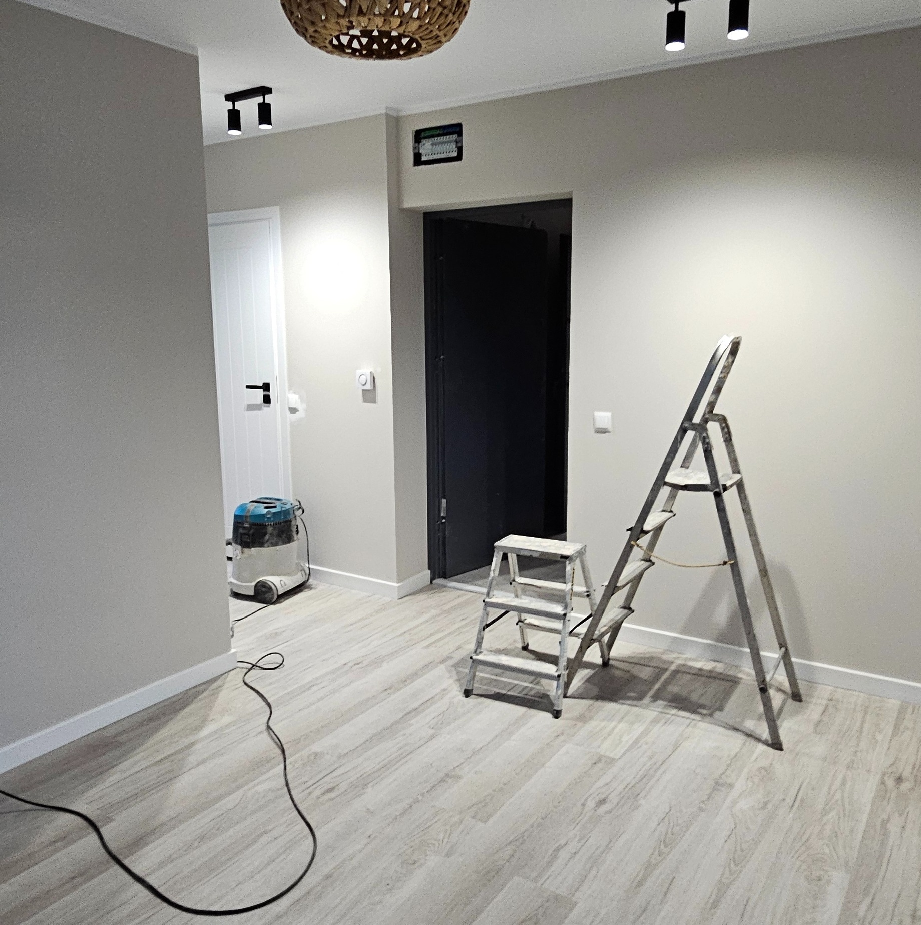 Jasne wnętrze po malowaniu ścian z drabiną, odkurzaczem i drewnianą podłogą. Minimalistyczny design, neutralna kolorystyka, oświetlenie punktowe, świeżo po remoncie.