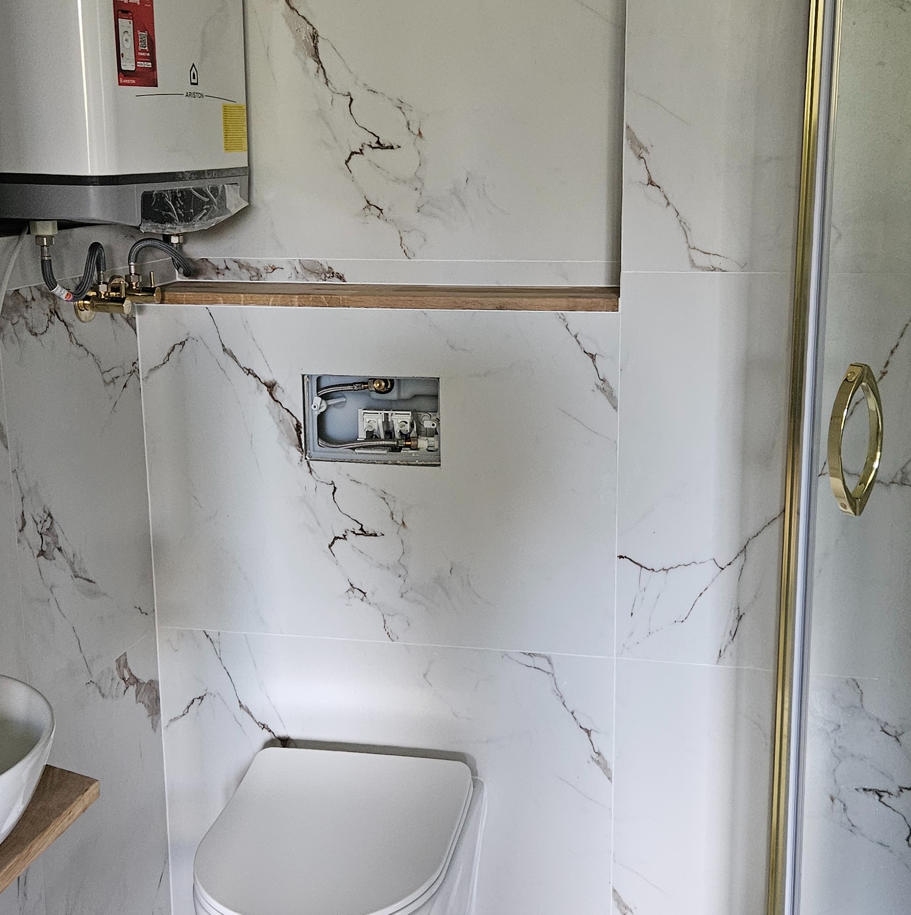 Biała łazienka z marmurowymi płytkami, widoczny podtynkowy stelaż WC, bojler i kabina prysznicowa z złotymi detalami. Minimalistyczny design i eleganckie wykończenie.