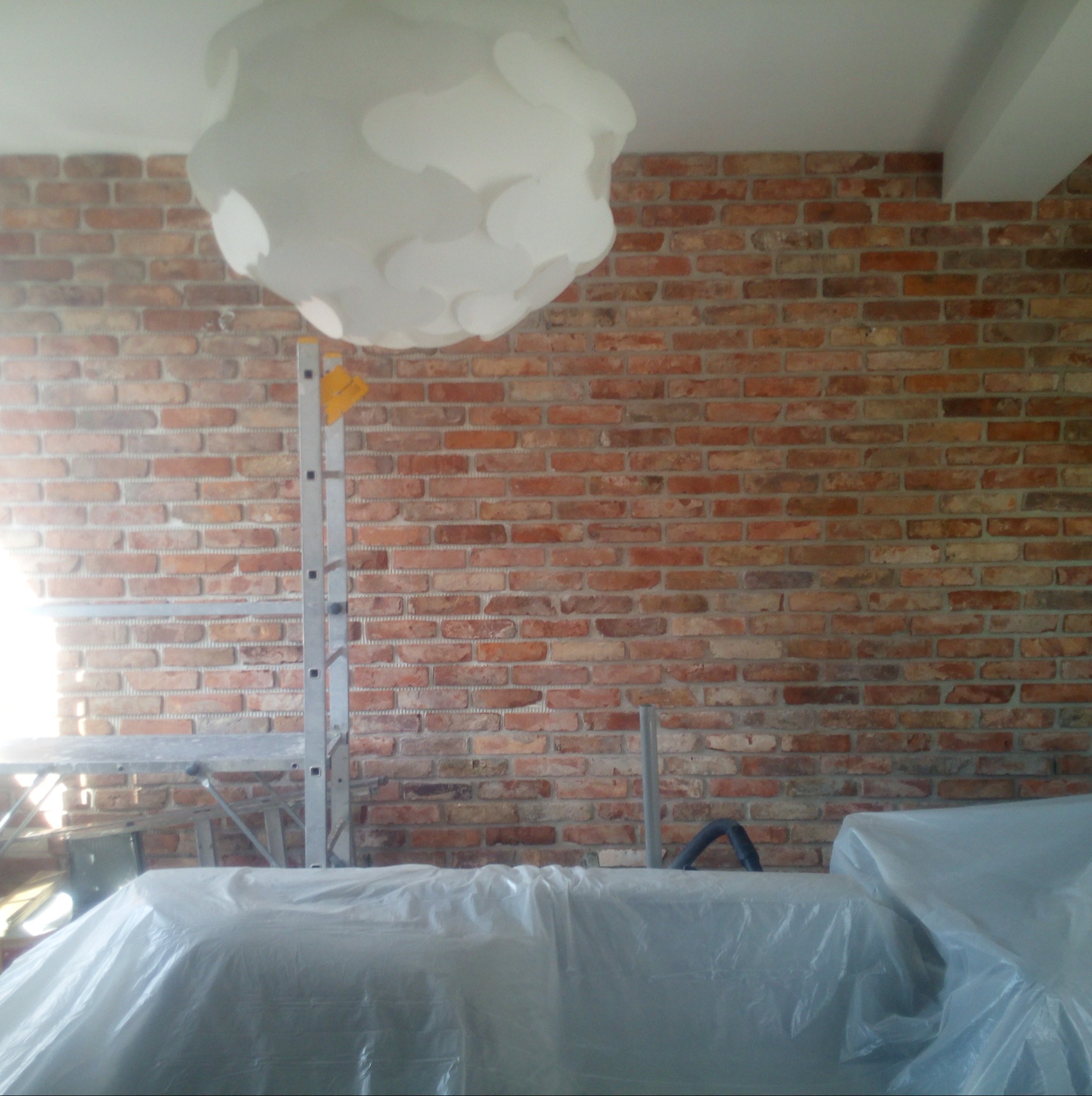 Murowana ściana z cegły po renowacji, zabezpieczona folią malarską, widoczna drabina i lampa sufitowa. Detal wnętrza po pracach wykończeniowych.