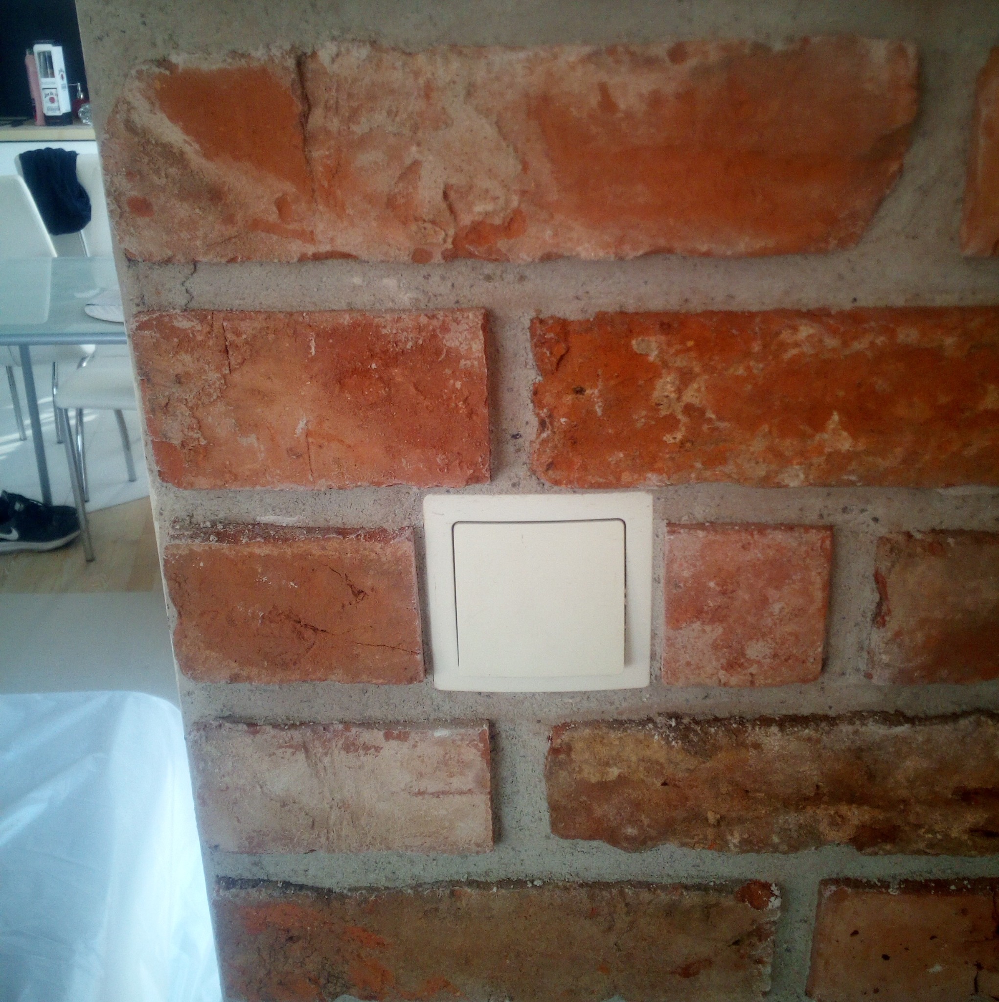 Ściana z cegły z włącznikiem światła. Widoczne fragmenty mebli w tle. Nietypowe wykończenie ściany z dekoracyjną cegłą i osprzętem elektrycznym.