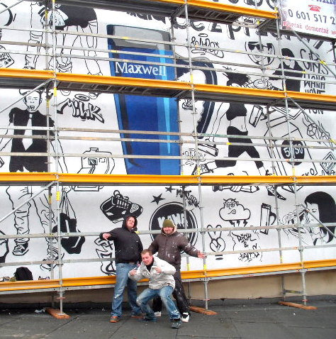 Ekipa pozuje na tle muralu z rysunkami i reklamą Maxwell, ustawionego na rusztowaniu. Czarno-biała grafika z elementami koloru niebieskiego.