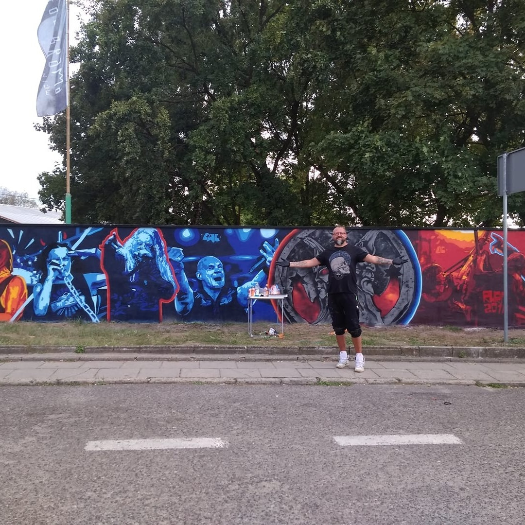 Artysta prezentuje mural z wizerunkami muzyków, wykonany na betonowym murze przy ulicy. Na stoliku farby w sprayu. Widoczne drzewa i flaga.