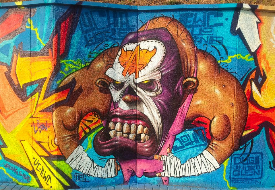 Graffiti na ścianie: kolorowy mural z karykaturalną postacią z odsłoniętymi zębami i literą 'A' na czole, otoczoną abstrakcyjnymi wzorami i napisami.