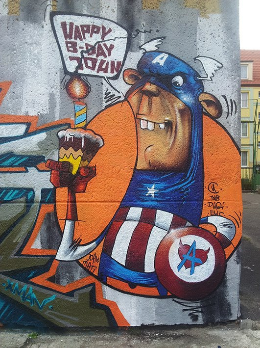 Humorystyczny mural na ścianie: karykatura Kapitana Ameryki z tortem urodzinowym i napisem 'Happy B-Day John'. Graffiti w stylu street art z żywymi kolorami.