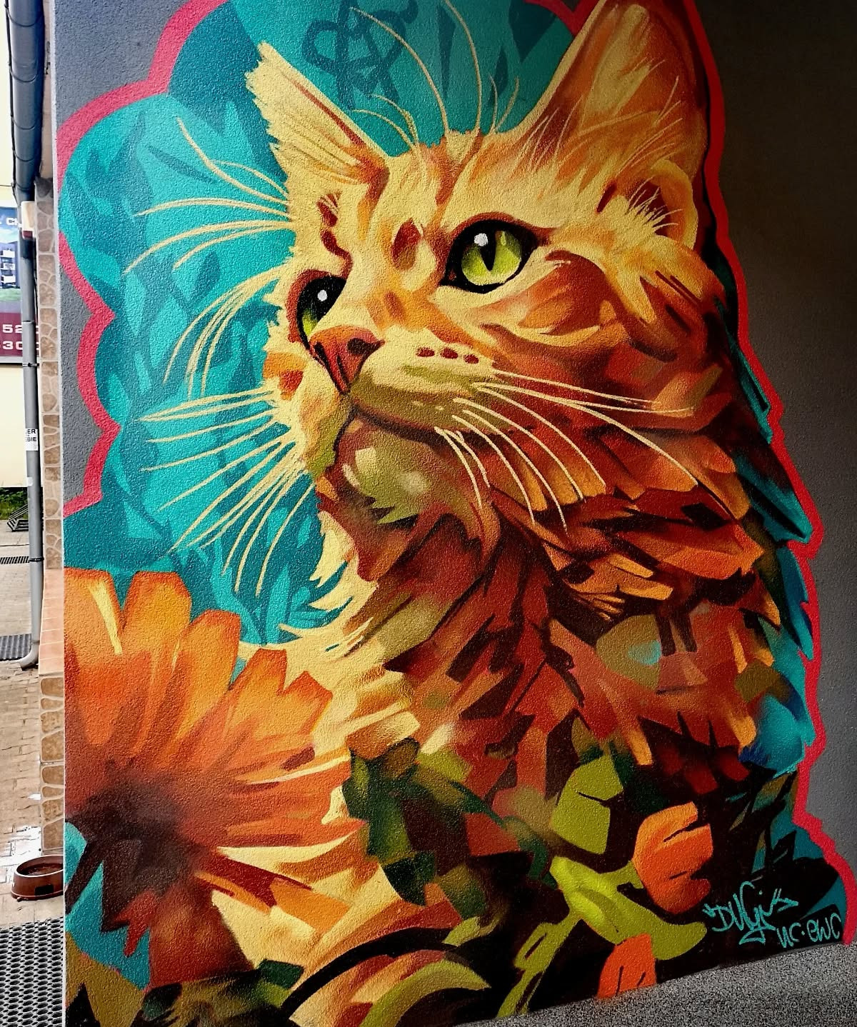 Artystyczny mural z wizerunkiem rudego kota z zielonymi oczami na tle błękitnych wzorów i pomarańczowych kwiatów, wykonany na szarej ścianie budynku.