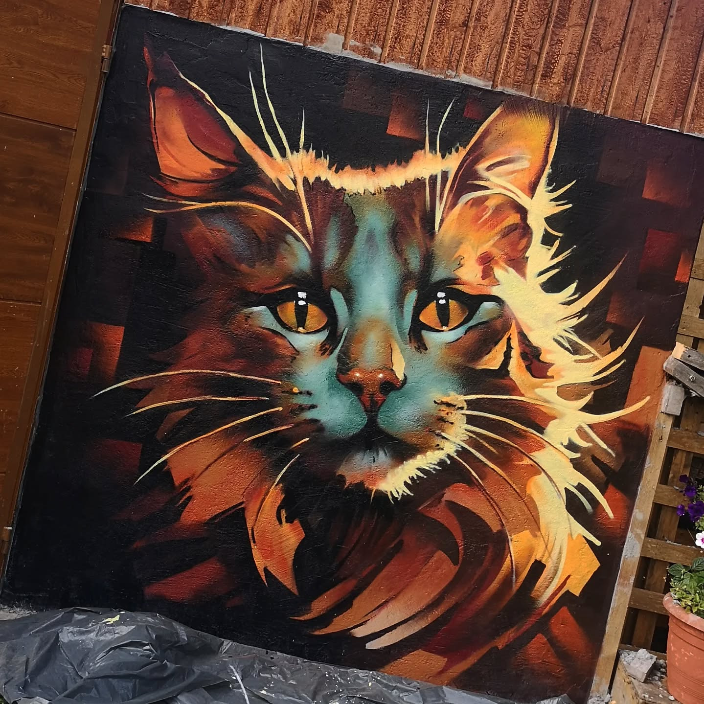 Portret kota w stylu pop-art na płótnie, z intensywnymi pomarańczowymi i turkusowymi akcentami, ustawiony na zewnątrz przy drewnianym ogrodzeniu.