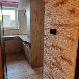 Albedo - Wykończona kuchnia z drewnianą podłogą i ścianą z dekoracyjnej cegły. Widoczne meble kuchenne i okno z fioletowym oświetleniem LED.