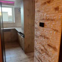 Wykończona kuchnia z drewnianą podłogą i ścianą z dekoracyjnej cegły. Widoczne meble kuchenne i okno z fioletowym oświetleniem LED.