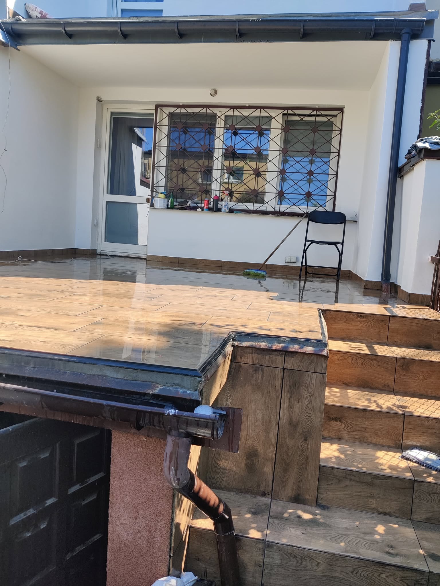 Błyszcząca, świeżo umyta podłoga na tarasie z drewnopodobnych płytek. Widoczne schody i okno z kratą. Na podłodze leży mop.