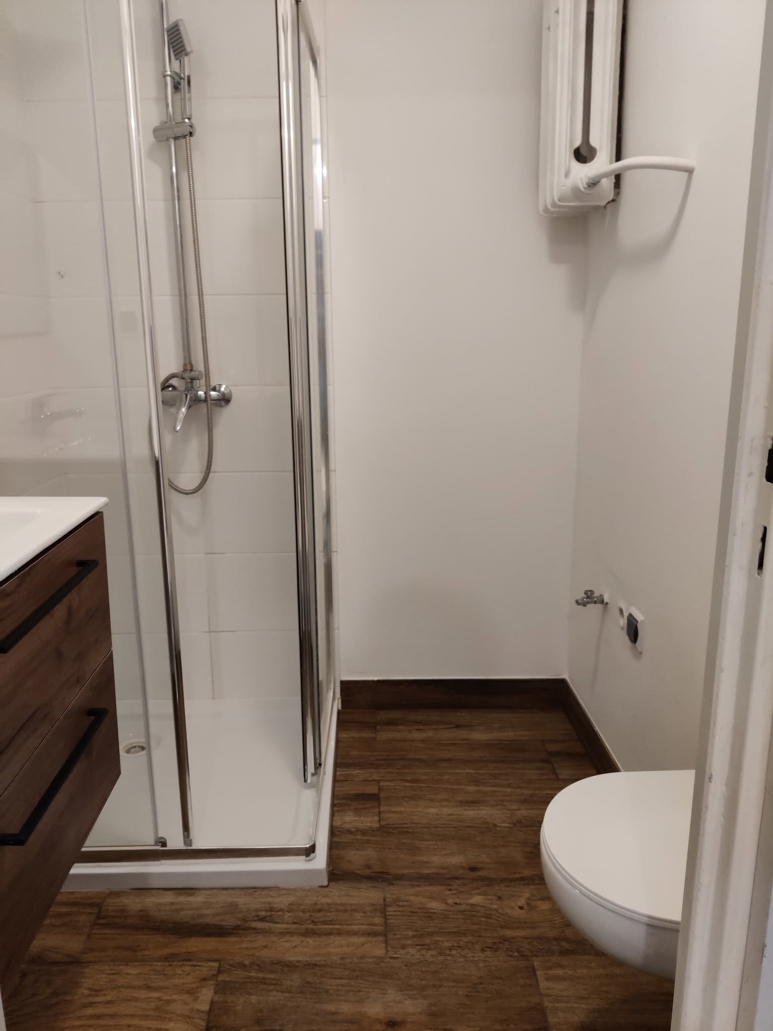 Nowoczesna łazienka po remoncie: kabina prysznicowa ze srebrnymi okuciami, drewnopodobne panele podłogowe i biała armatura. Grzejnik żeberkowy na ścianie.