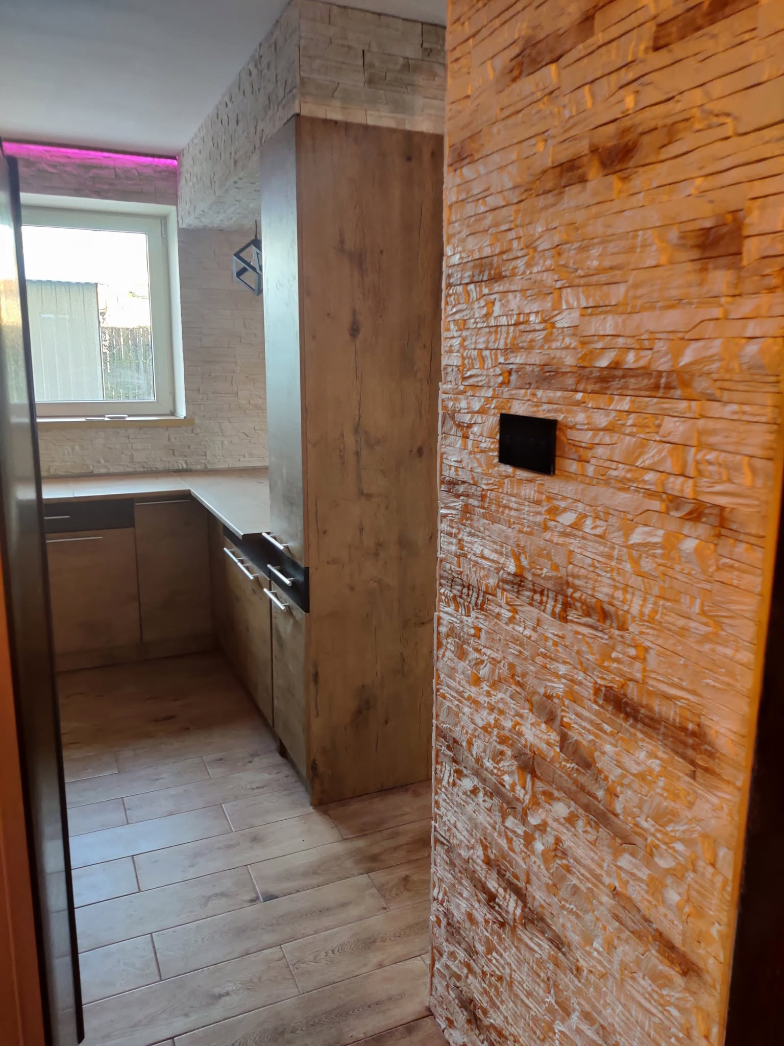 Wykończona kuchnia z drewnianą podłogą i ścianą z dekoracyjnej cegły. Widoczne meble kuchenne i okno z fioletowym oświetleniem LED.