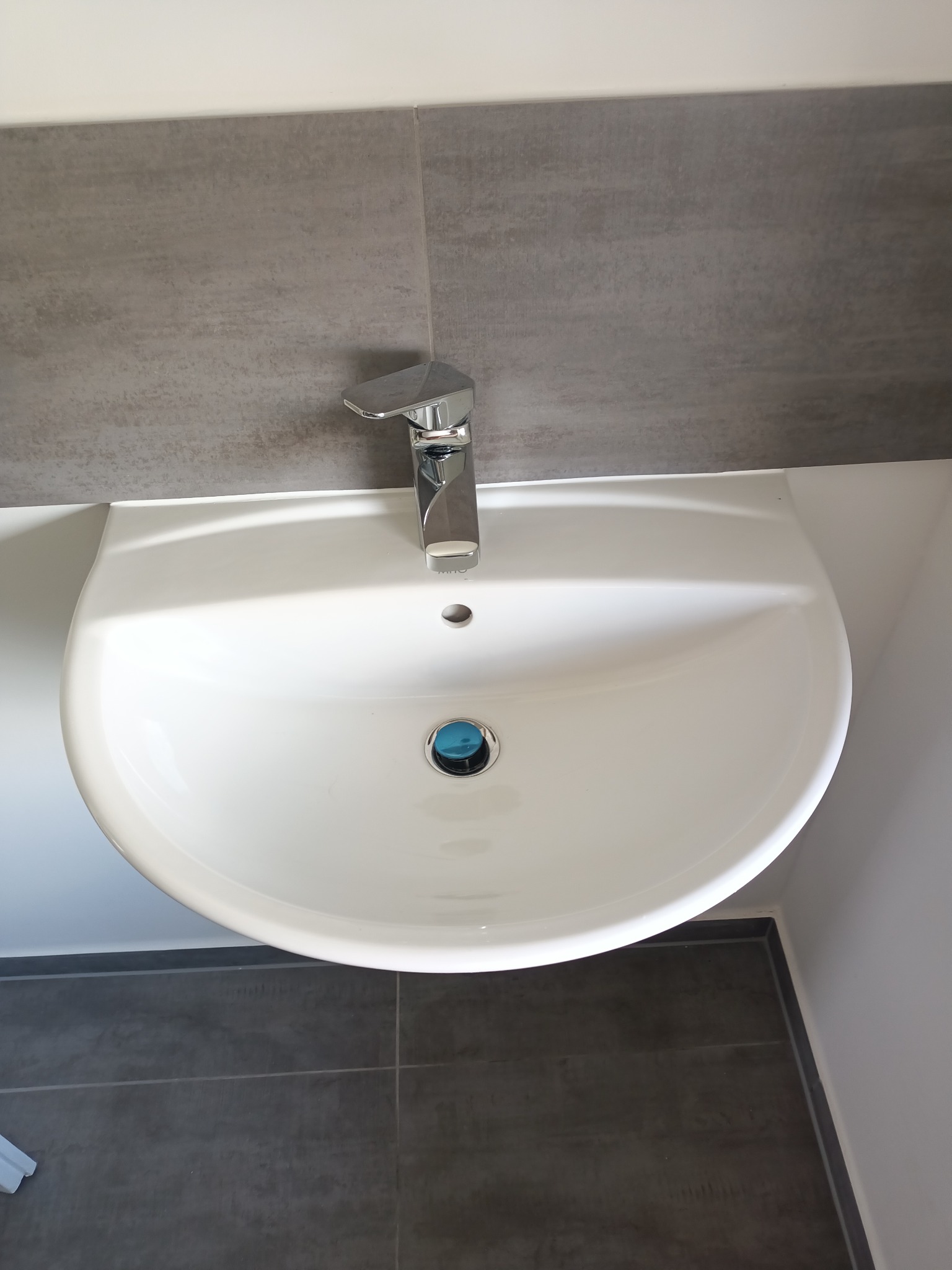 Biała umywalka z chromowaną baterią na tle szarych płytek ściennych i podłogowych. Minimalistyczny design łazienki, widok z góry.